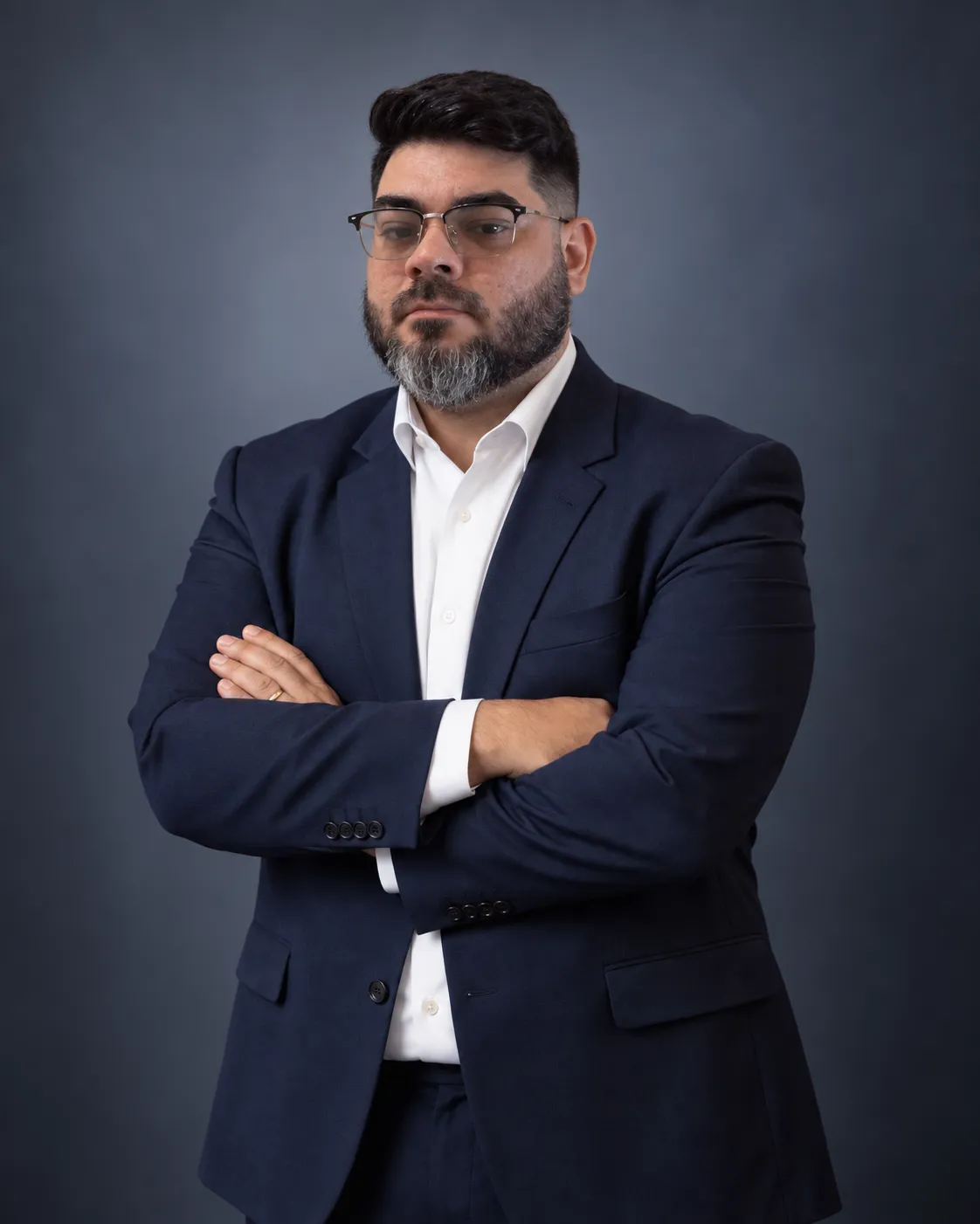 Gabriel Walraven em pé, de braços cruzados, com terno azul-marinho sobre fundo em gradiente cinza